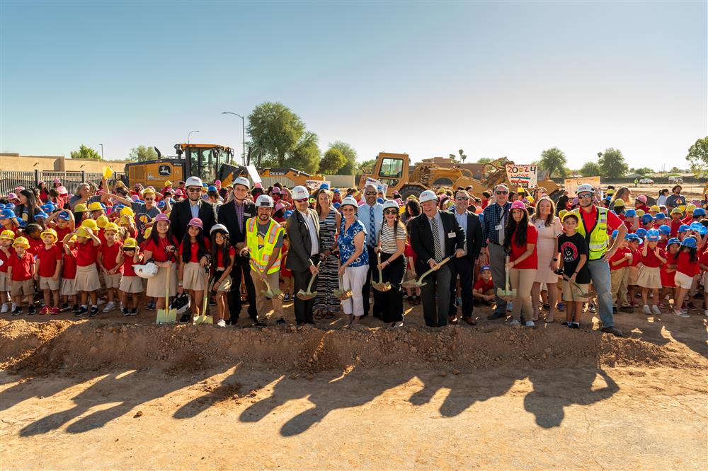 People dressed in hard hats and safety vests use shovels to dig into the ground